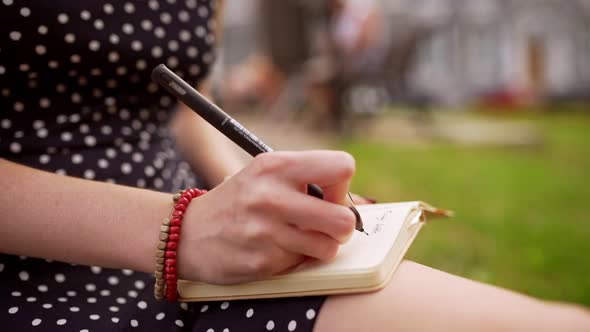 Girl Writing Notes with Pen While Sitting in the Street in Polka Dot Dress with Naked Legs and Arms