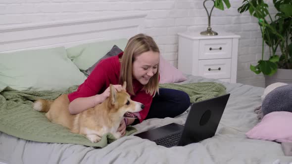 Teenage Girl with Funny Corgi Dog and Laptop on Bed at Home