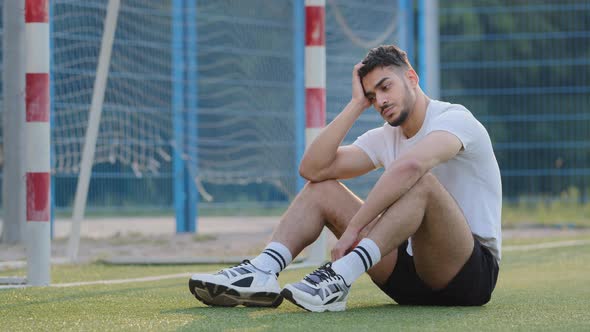 Frustrated Sad Middle Eastern Footballer Sitting on Grass of Soccer Field Against Goal Worried