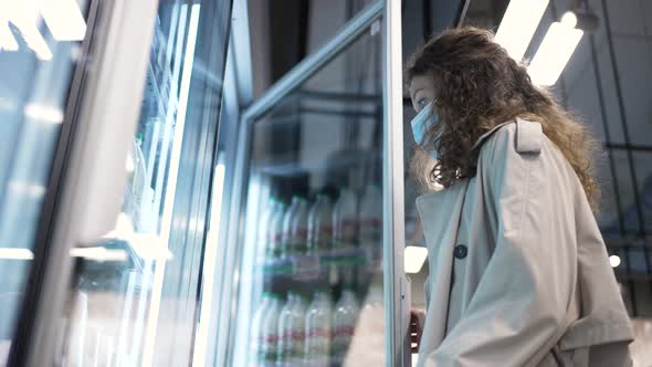 Woman Shopper in a Medical Protective Mask Opens a Large Refrigerator in a Supermarket and Chooses