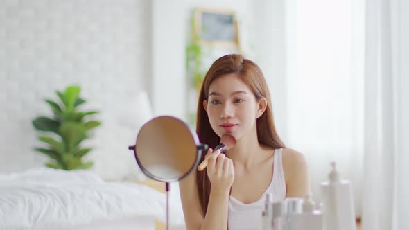 Happy Asian young woman smile and looking in mirror doing skin care applying make up brush on cheek