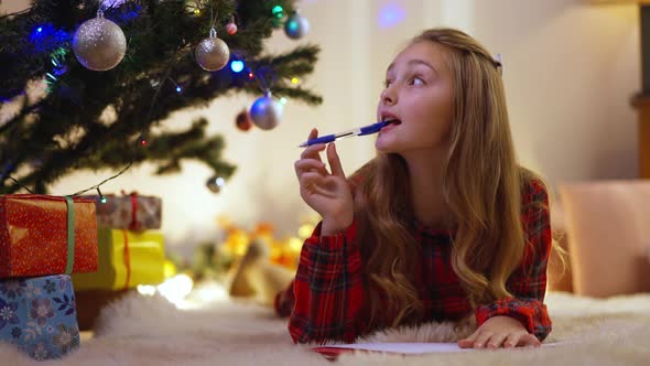 Cute Little Girl Writing Letter to Santa Lying on Soft Carpet Under Christmas Tree at Home