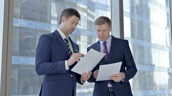 Two Middle Aged Businessmen Reading Documents Against Boardroom Office Window