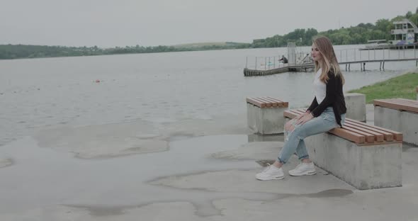 Girl Sitting on Bench at Riverside and Admiring the Waves in Windy Weather