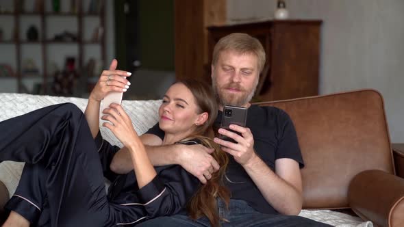 Spouses Are Embracing at Couch in Home and Using Smartphones, Man and Woman