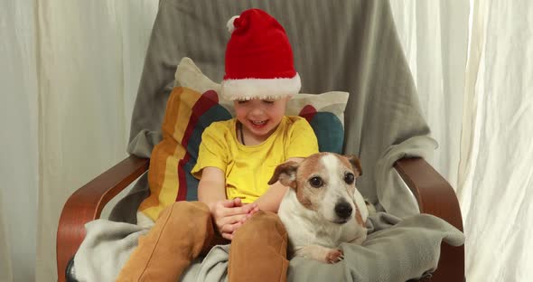 Young Boy in Christmas Hat and T-shirt Pets Dog on Chair