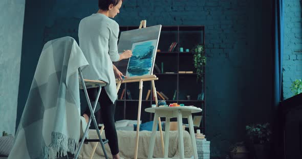 Young Woman Paints a Landscape on Canvas in a Bright Workshop