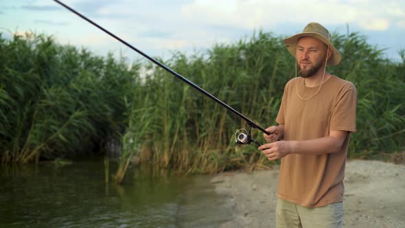 a Man Fisherman at Sunset in a Hat on the Lake Catches Fish with a Fishing Rod Against the