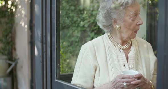 Senior Grandma Woman Enjoying Drinking Cup of Tea or Coffee Near Window Looking Outside