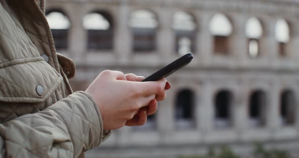 A Woman Dials a Message on Her Mobile Phone While Standing in Front of Coliseum