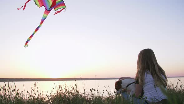 Young Happy Woman and Het Little Dog Sitting with Flying Kite on a Glade at Sunset