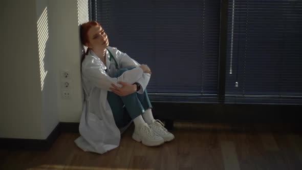 Frustrated Young Female Doctor in White Coat Sitting on Floor Near Window with Louvre in Dark