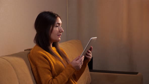 Concentrated Brunette in Yellow Sweater Reads Ebook on Sofa