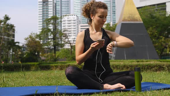 Young Woman Doing Yoga Using Smartphone And Smartwatch At City Park