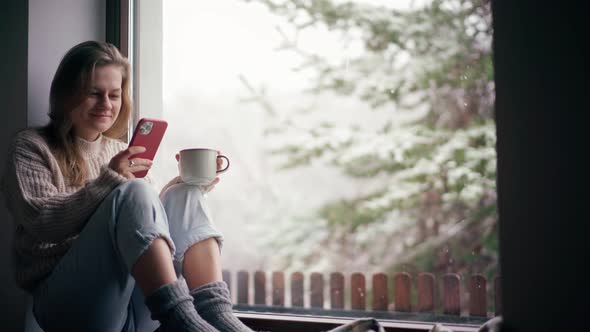 A Young Girl Sits on the Windowsill and Drinks Coffee While Using Her Smartphone