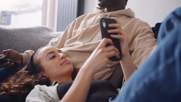 Relaxed Young Couple At Home Sitting On Sofa Watching TV And Checking Social Media On Mobile Phone