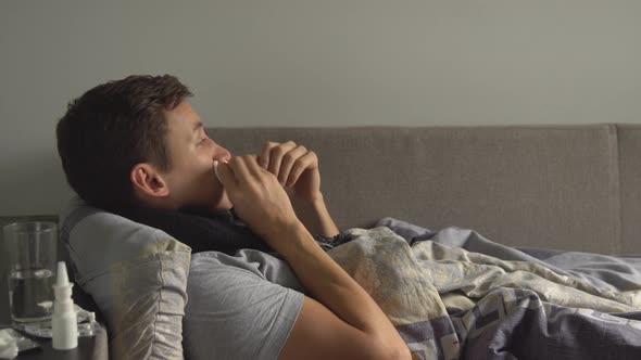 Man Lying Sick in Bed Next To His Medications in His Home and Blowing His Nose in a Handkerchief