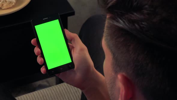 A Man Watches Something on a Smartphone with a Green Screen in Living Room