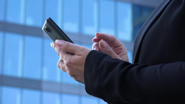 A Middle-aged Businesswoman Works on a Smartphone - Closeup - a Windowed Office Building