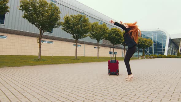 Happy Young Woman Dancing Near Luggage Suitcase on Airport Terminal Background. A Woman with Red