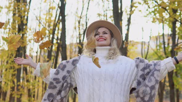 Beautiful Blonde Woman in a Brown Hat Throws Leaves in the Autumn Park