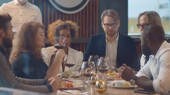 Waiter in Protective Face Shield and Mask Serving Food for Diverse Group of People in Restaurant