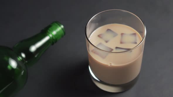 Ice cubes pop up in cream Irish liquor in a glass on a dark table with an empty green bottle. 