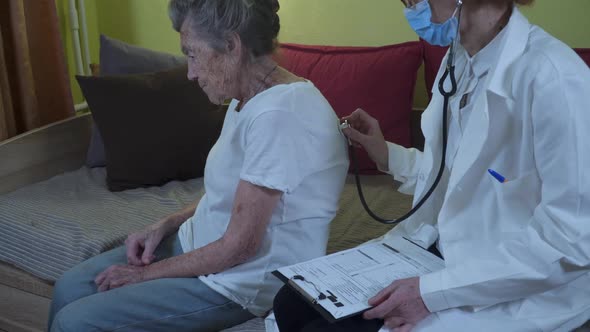 Mature Woman Doctor Holding Stethoscope Examining Senior Grandma Patient at Home