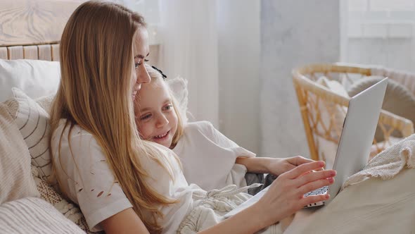 Mom and Daughter Lie in Bed Looking in Laptop