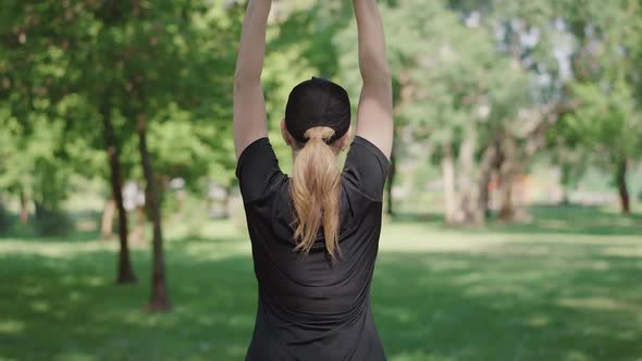 Back View of Blond Mid-adult Sportswoman in Black Cap and T-shirt Raising Hands Up and Bending. Slim