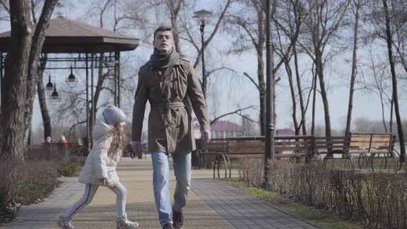 Joyful Little Girl Running and Jumping Around Young Caucasian Man. Tired Father Walking with