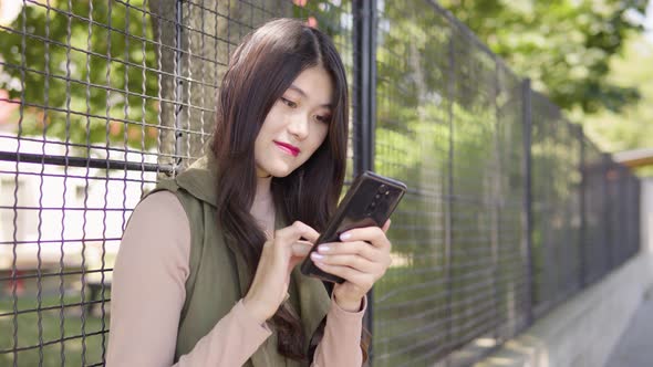 Young Asian Woman Works Smartphone Smile As She Leans Against Tall Fence