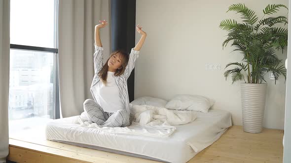Young Chinese Woman Dressed in Pajamas Stretching Waking Up Sitting on Her Bed on Her Day Off