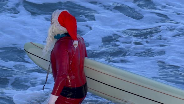 Santa Claus walking with his surfboard to go surfing at the beach.