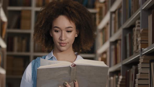 Young African American Girl Student Stands in Public University Library Interested in Reads Book