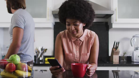 African american woman holding coffee cup using smartphone in the kitchen at home