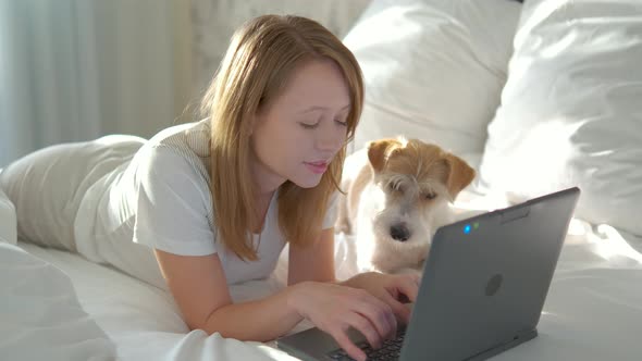 A beautiful girl in casual clothes is lying on the bed and typing on a laptop.