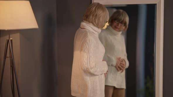 Senior Woman Looking in Mirror Standing in Living Room at Home Indoors