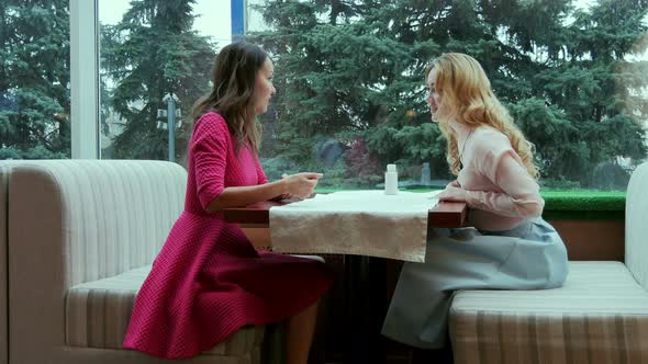 Two Smiling Beautiful Young Women Talking at the Table in Cafe