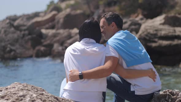 Romantic couple sit and embrace near the sea. Man and woman with black hair spend vacation