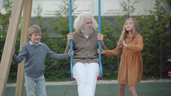 Front View of Relaxed Senior Caucasian Man Sitting on Swings As Boy and Girl Talking and Smiling