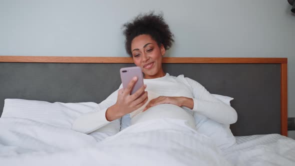 Positive African pregnant woman looking at phone on the bed
