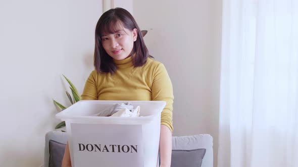Portrait of young asian woman looking at camera holding box with clothes for donation.