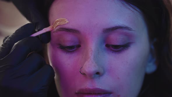 A Makeup Artist Waxing the Eyebrow Hairs of His Female Client in Neon Lighting