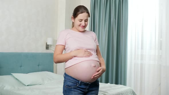 Smiling Pregnant Woman Caressing and Touching Big Belly While Standing in Bedroom with Big Window