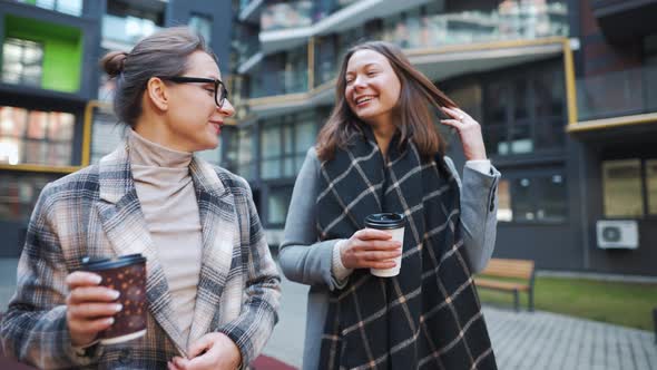 Two Happy Women Walking with Takeaway Coffee and Talking with Interest Among Themselves in the