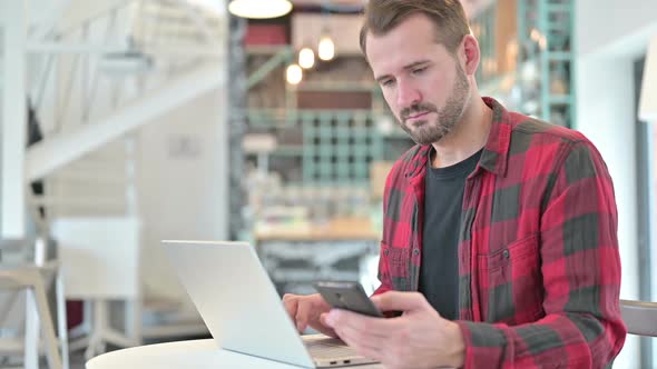 Young Man with Laptop Using Smartphone in Cafe