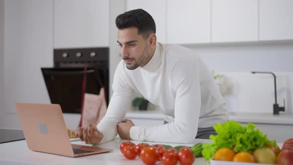 Concentrated Middle Eastern Young Man Surfing Internet on Laptop Standing at Countertop with