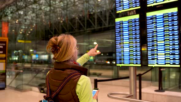 Blond woman points to flight information display while holding her smartphone in an airport.