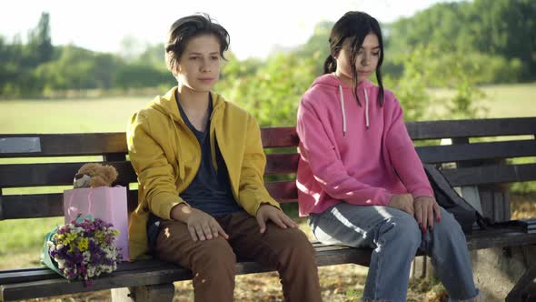 Portrait of Shy Teenage Boyfriend Sitting with Girlfriend in Summer Park As Girl Putting Palm on